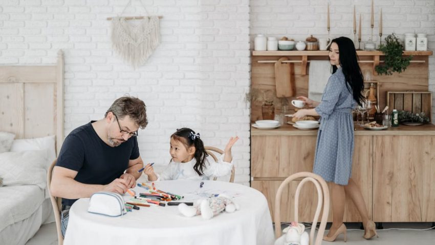 Father and daughter bond over crafts while mother prepares food in a cozy home setting.